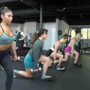 Four female clients performing reverse lunges in a straight line during a glute workshop at Fitness Factory KC. Each participant is focused, demonstrating proper form with their hands on their hips, engaging their glutes and leg muscles.